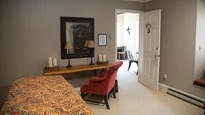 Un dormitorio con una cama, una silla roja, un escritorio con una lámpara y una puerta que da a otra habitación.