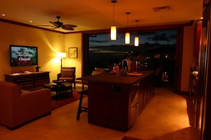 A cozy living room with a TV, comfortable seating, a kitchen area, and a large window with a view. - Oceanfront 2BR 2BA Villa Top-Floor Corner Walk to Disney Aulani Sleeps 6 (Kapolei)