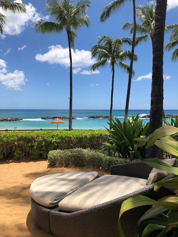A beach view with palm trees, a wicker lounge chair, and a thatched umbrella. - Oceanfront 2BR 2BA Villa Top-Floor Corner Walk to Disney Aulani Sleeps 6 (Kapolei)
