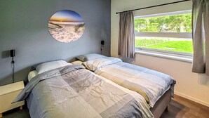 Een slaapkamer met een bed, een nachtkastje, een rond kunstwerk aan de muur, een raam met gordijnen en uitzicht op groen.