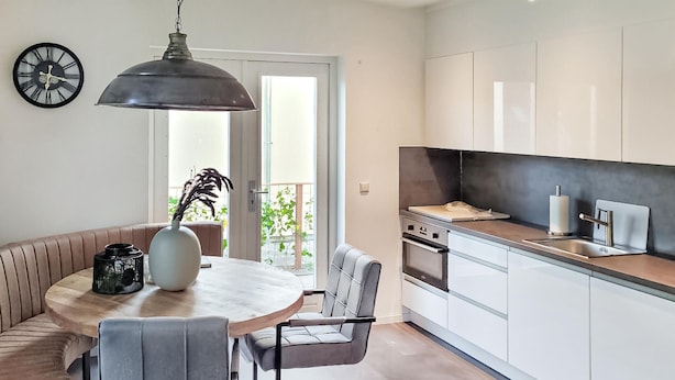 Een moderne keuken met witte kasten, een zwart aanrecht en roestvrijstalen apparatuur. Er is een eethoek met een ronde tafel en stoelen, en een groot raam zorgt voor natuurlijk licht.