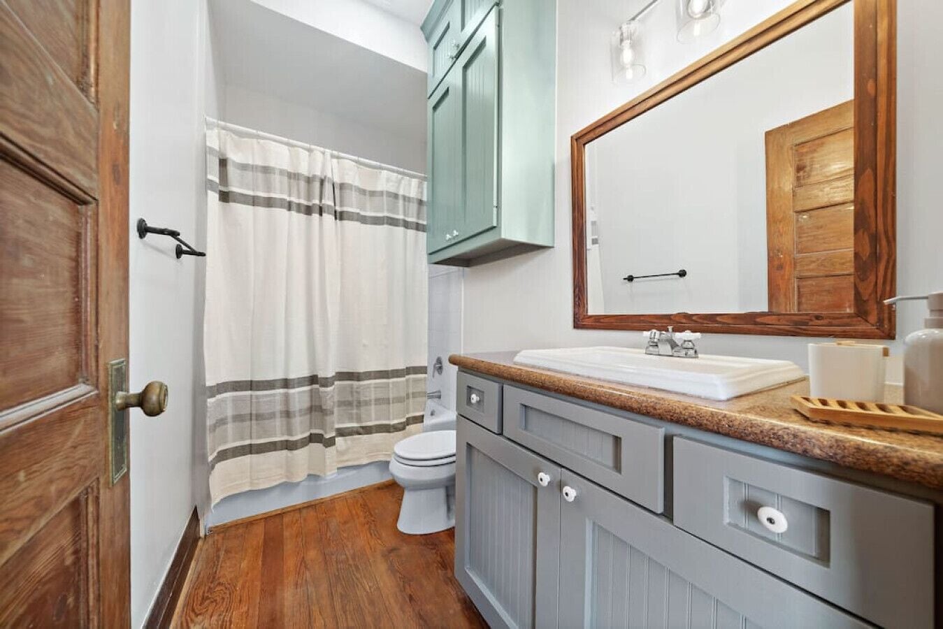 Un baño con piso de madera, un inodoro blanco, cortina de ducha a rayas y un tocador con espejo y lavamanos.