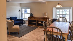A spacious living area with a blue sofa, a wooden dining table, and chairs. There is a lamp on the table and a window with a view of the outside. - 3 bedroom accommodation in Swanage (Swanage)
