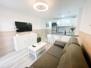 A modern living room with a sofa, a coffee table, and a TV. There is a kitchen area with a dining table and chairs. - Pool View Holiday Home (Playa del Inglés)