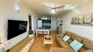 Ein modernes Wohnzimmer mit Fernseher, einem beigen Sofa und einem Essbereich mit weißem Tisch und Stühlen.