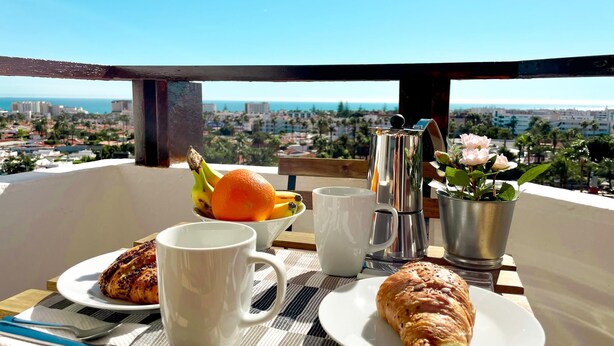 Frühstücksaufteilung mit Croissants, einer Kaffeekanne und einer Obstschale auf einem Balkon mit Blick auf eine Stadtlandschaft.