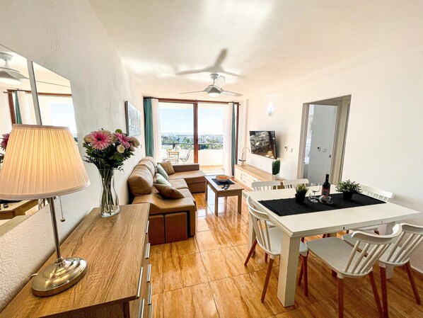 A modern living room with a sofa, dining table, and chairs. There is a ceiling fan, a TV, and a view of the city through large windows.
