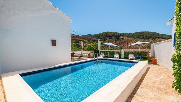 A rectangular swimming pool with clear blue water, surrounded by a white wall and a stone patio. There are lounge chairs and umbrellas in the background, with a view of green hills under a clear sky. - La Casa de Aulabar Belmez de la Moraleda by Ruralidays (Jaén)