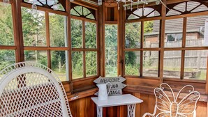 A wooden structure with large windows, a white wicker chair, a small table with a sign, and a decorative metal chair.