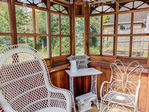 A wooden structure with large windows, a white wicker chair, a small table with a sign, and a decorative metal chair.