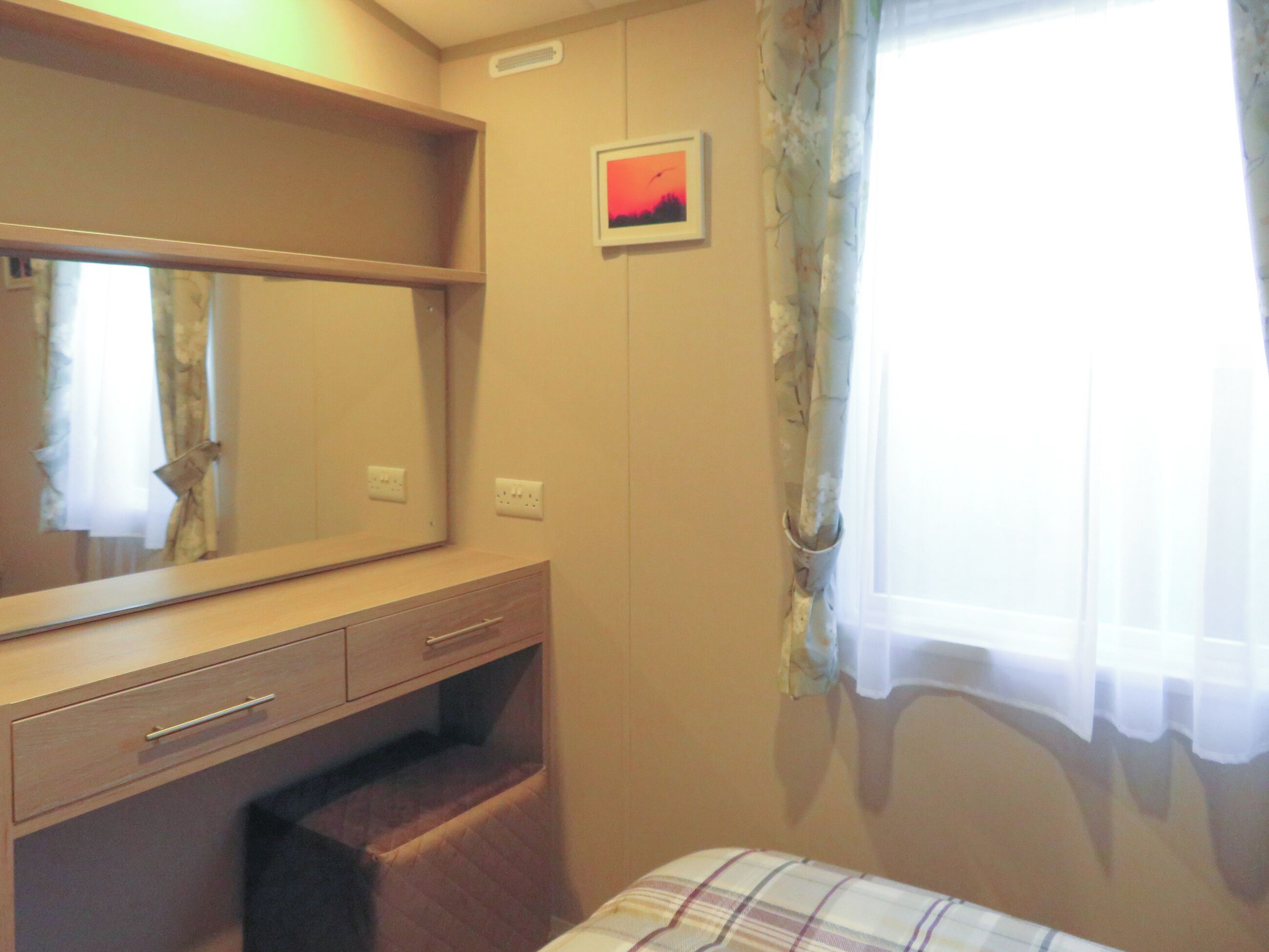A bedroom with a bed, a bedside table, a mirror, a small picture on the wall, and a window with curtains.