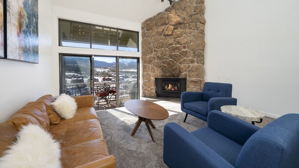 A modern living room with a stone fireplace, blue sofas, and a large window overlooking a snowy landscape. - Spacious, Updated Mountain Home with Views, Private Hot Tub and Sauna. (Fraser)