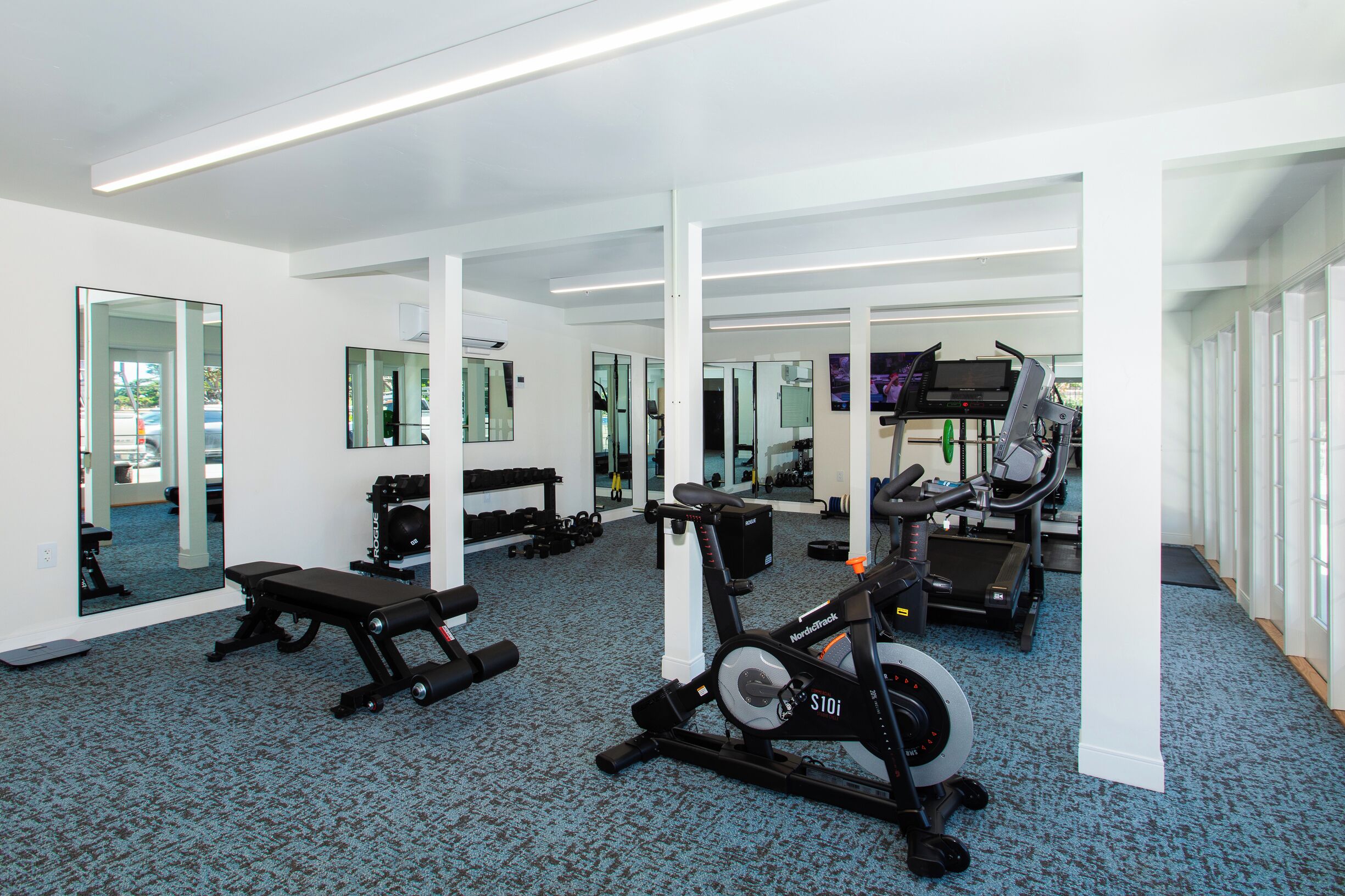 A modern gym with mirrored walls, exercise equipment, and a blue carpeted floor.