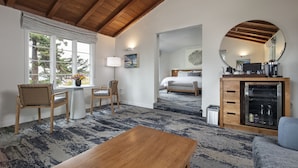 A hotel room with a wooden ceiling, a large window with a view of trees, a bed, a desk with a chair, a round mirror, and a cabinet with kitchen appliances.