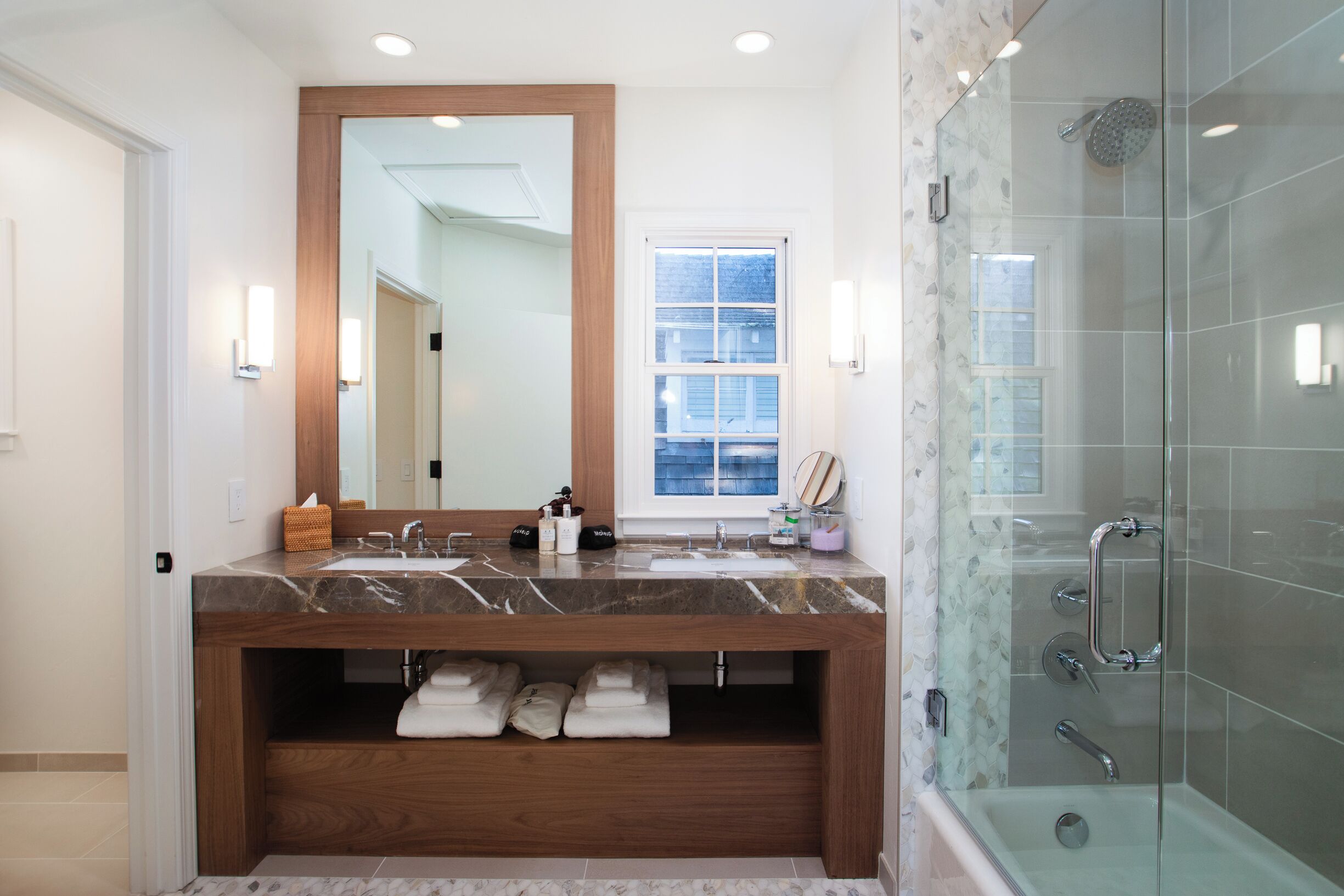 A modern bathroom with a marble sink, wooden vanity, glass shower enclosure, and a large mirror.