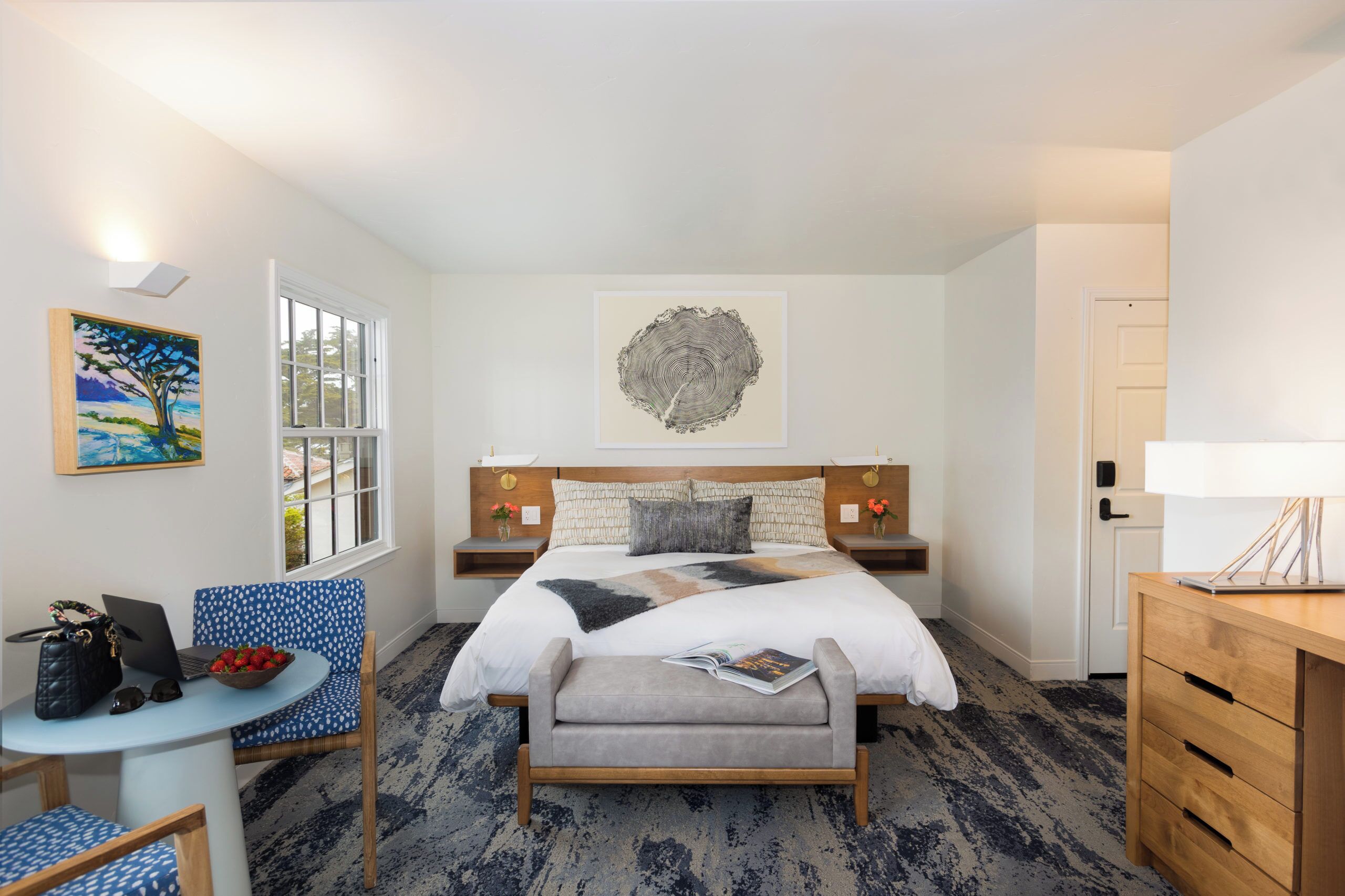 A hotel room with a bed, a desk with a laptop, a chair, a small table with a bowl of fruit, and artwork on the walls.