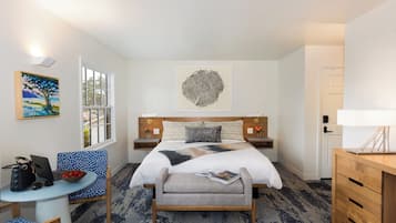 A hotel room with a bed, a desk with a laptop, a chair, a small table with a bowl of fruit, and artwork on the walls.