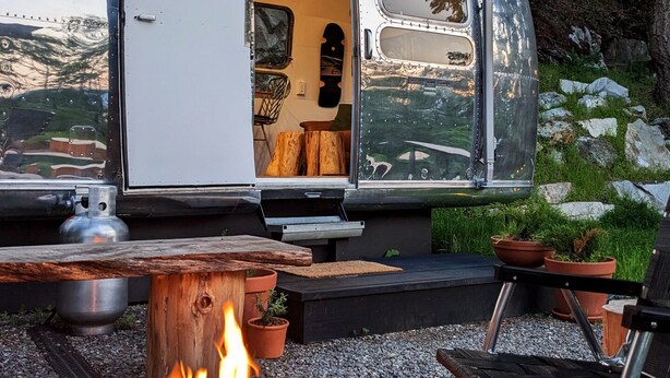 A camper with an open door revealing a kitchen area, a fire pit, and outdoor seating. - Wildernest Mountainside Airstream Getaway (Bowen Island)