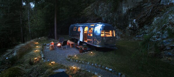 Una autocaravana con un brasero y zona de estar rodeada de bosque.