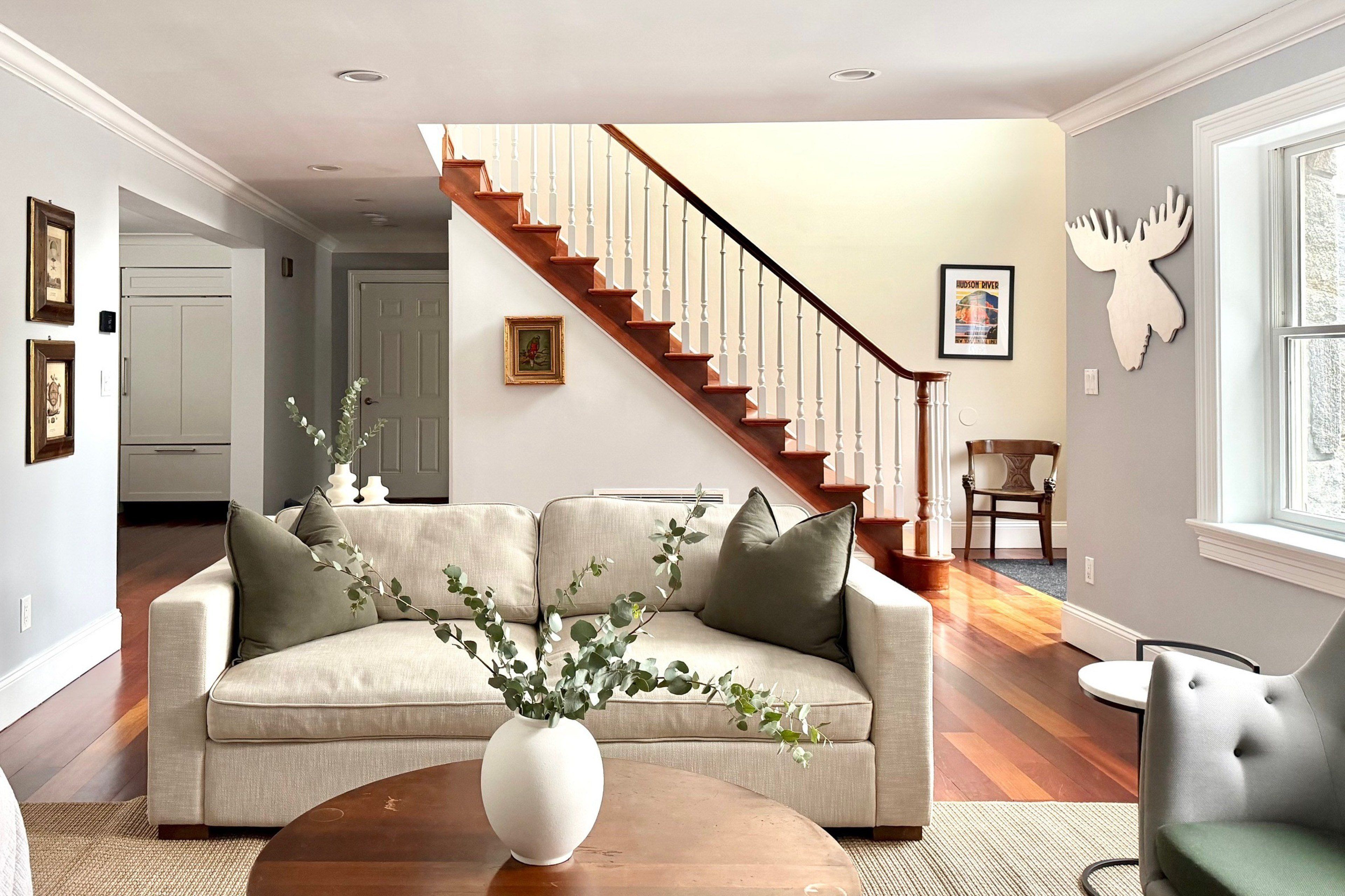Un salon avec un canapé beige, une table basse ronde en bois et un escalier avec des marches en bois et des balustrades blanches.