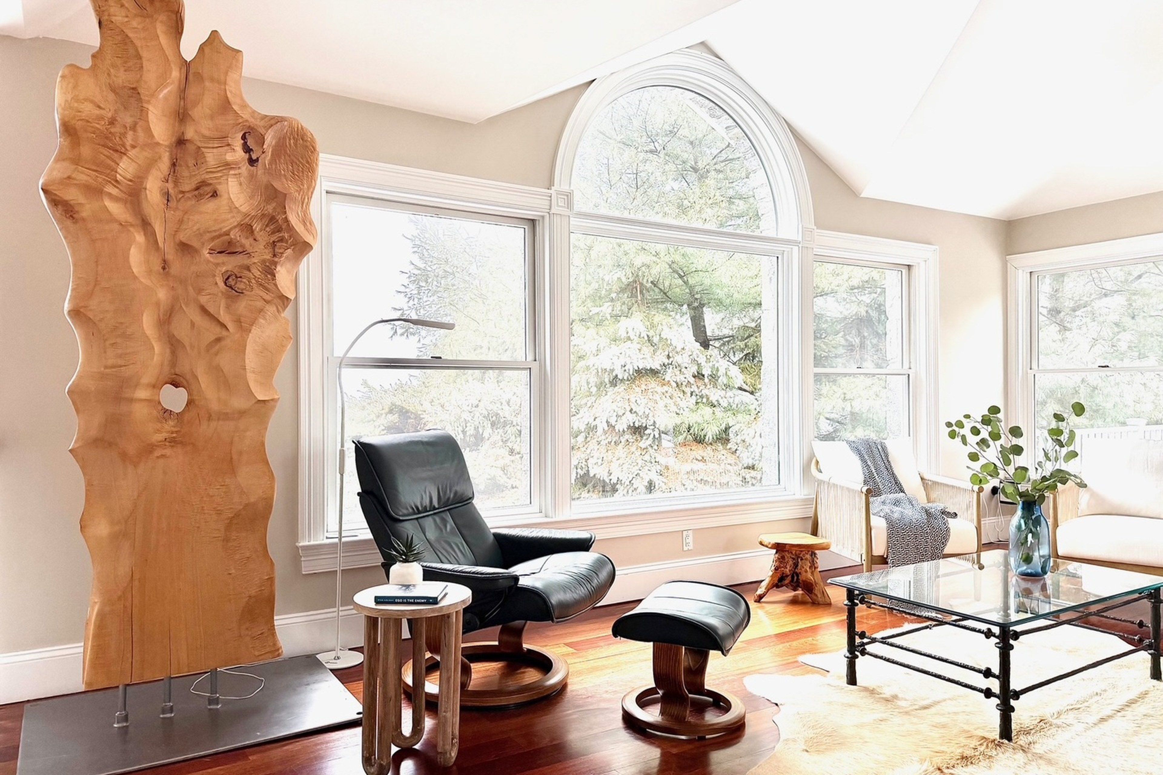 Una sala de estar moderna con una gran escultura de madera, sillones de cuero, una mesa de centro de vidrio y ventanas amplias con vistas a árboles.
