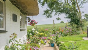 A white cottage with a thatched roof, surrounded by a well-maintained garden with various flowers and plants. - Brittons Hill Cottage (Kenardington, near Ashford)