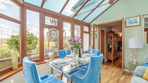 A conservatory with a glass table set for dining, blue upholstered chairs, and a view of the countryside. - Brittons Hill Cottage (Kenardington, near Ashford)