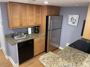 A compact kitchen with wooden cabinets, a stainless steel refrigerator, a dishwasher, and a granite countertop.
