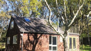A small, single-story wooden house with a sloped roof, a large window, and a small porch area with a swing chair.