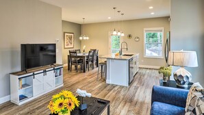 A modern kitchen with a central island, a dining area with dark wooden chairs, a built-in TV, and a cozy seating area with a blue sofa. - Modern Candler Home w/ Deck & Yard! (Candler)