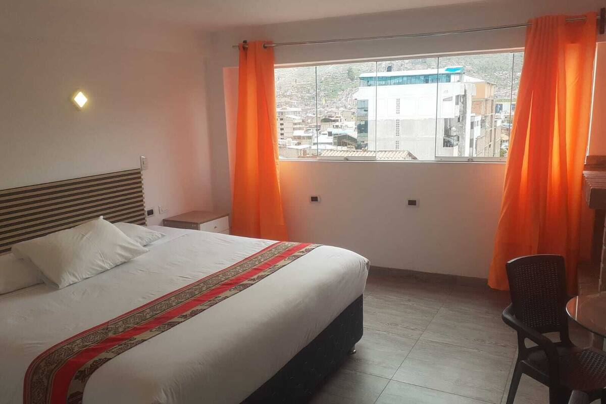 A hotel room with a bed, a chair, a window with curtains, and a view of buildings outside.