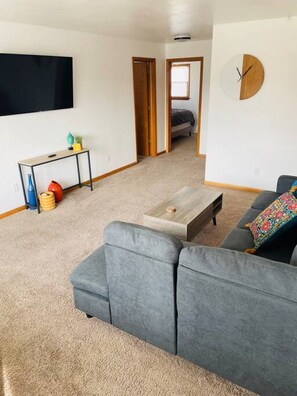 A living room with a grey sofa, a coffee table, a TV, and a door leading to another room. - Bay Street Unit 3 · Charming & Cozy APT-Free Parking (Appleton)