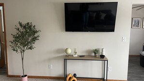 A living room with a TV mounted on the wall, a small console table with decorative items, a potted plant, and a pair of yellow slippers on the floor. - Chic & Cozy APT-Self Check-In (Appleton)