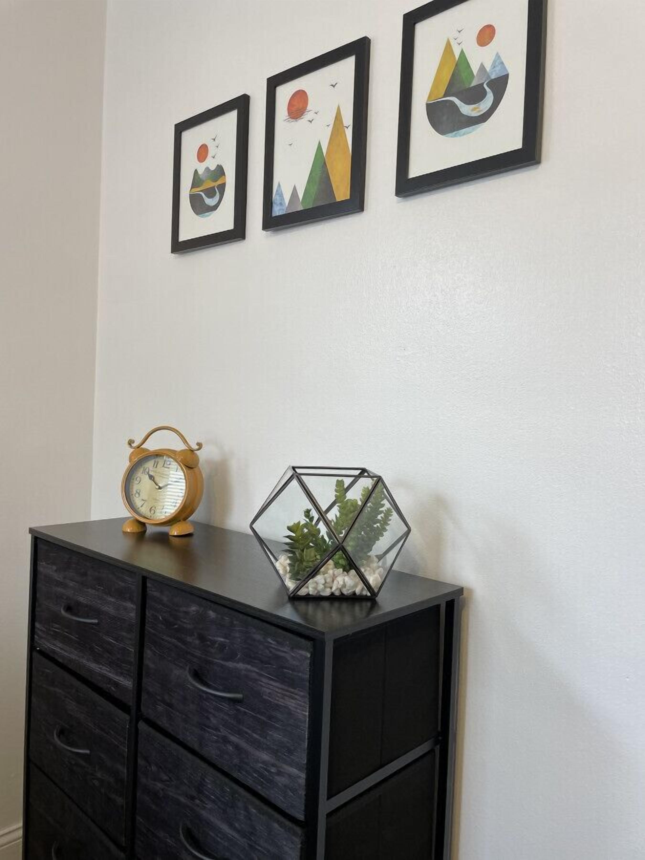 Een muur met daaraan drie ingelijste kunstwerken die abstracte landschappen voorstellen, een zwart metalen kast met lades, en een houten klok erbovenop.
