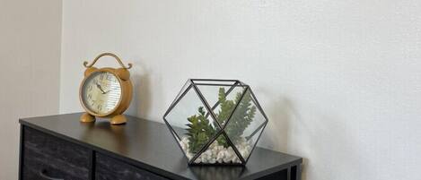 Een muur met daaraan drie ingelijste kunstwerken die abstracte landschappen voorstellen, een zwart metalen kast met lades, en een houten klok erbovenop.