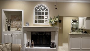 A living room with a fireplace, a large arched window, and a kitchen area with white cabinetry. - Farmhouse & Ranch style living for your vacay with pond, gazebo, trellis, pool.. (Farmersville)