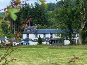 A large white building with a grey roof, surrounded by trees and a grassy field.
