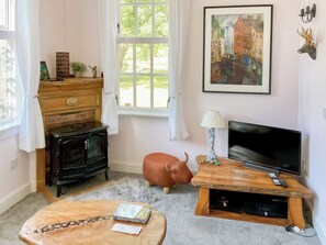 Ein gemütliches Wohnzimmer mit einem alten Kochherd, einem Fernseher, einem Holztisch und einem großen Fenster mit Vorhängen.