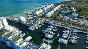 A marina with multiple boats docked, a row of white buildings, and a coastal view.