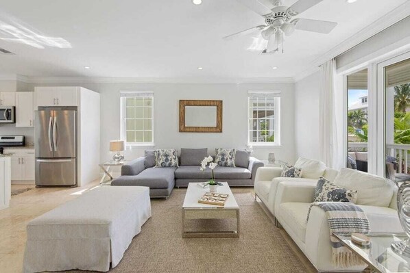 A modern living room with a grey sofa, a white sofa, and a coffee table. There is a ceiling fan, a microwave, and a refrigerator in the kitchen area.