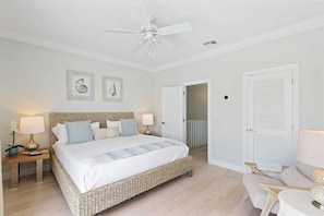 Um quarto com cama, mesas de cabeceira, ventilador de teto e uma porta que dá para outro cômodo.