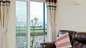 Un salon avec un canapé en cuir marron, un coussin à rayures et une vue sur une plage, avec des bâtiments blancs et une rambarde bleue.