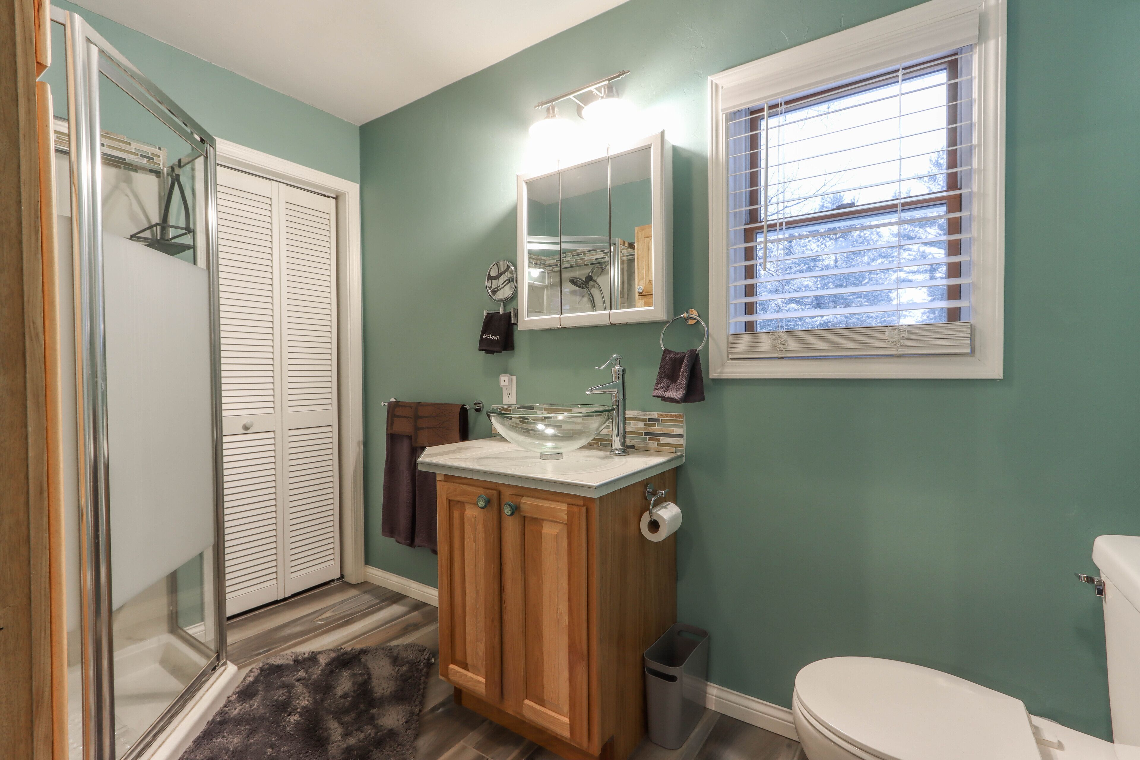 A bathroom with a glass sink, wooden cabinets, a white toilet, and a window with blinds.