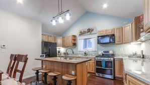 A modern kitchen with wooden cabinets, a central island with stools, a stainless steel refrigerator, a microwave, an oven, and a coffee machine.