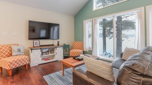 A living room with a TV, sofa, armchair, and a window with a view of trees.