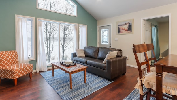 A living room with a brown leather sofa, a wooden coffee table, and a dining table with chairs. There are large windows with curtains, a ceiling fan, and a painting on the wall.