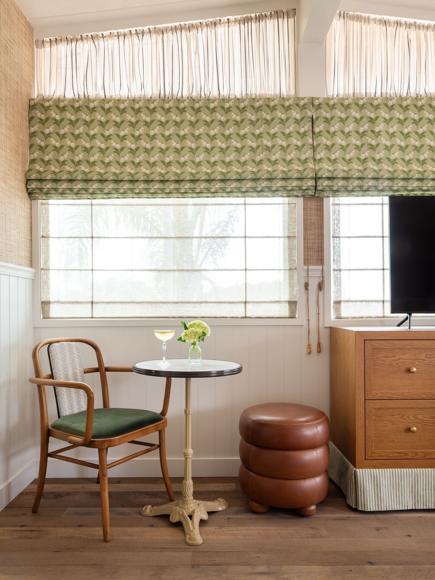 Una habitación con una silla de madera, una pequeña mesa redonda con un vaso, y un otoman marrón.