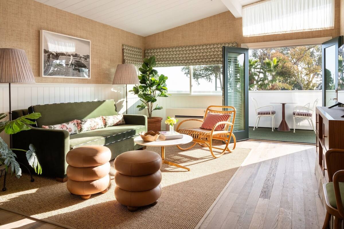 A modern living room with a green sofa, a round wooden coffee table, and a wicker chair. There is a large window with a view of trees and a balcony.