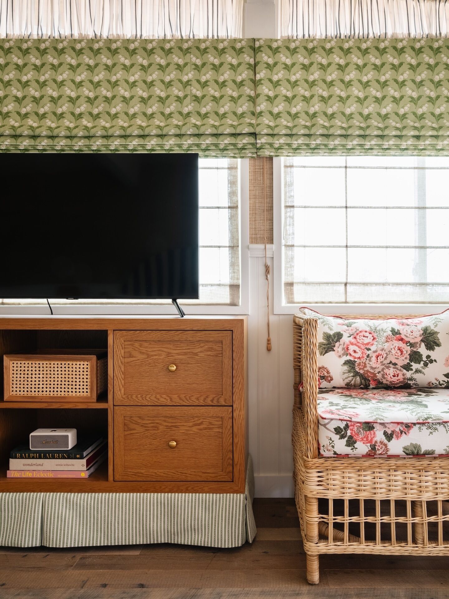 Een kamer met een televisie op een houten kast, een rieten stoel met bloemenstoffering, en een raam met dunne gordijnen.