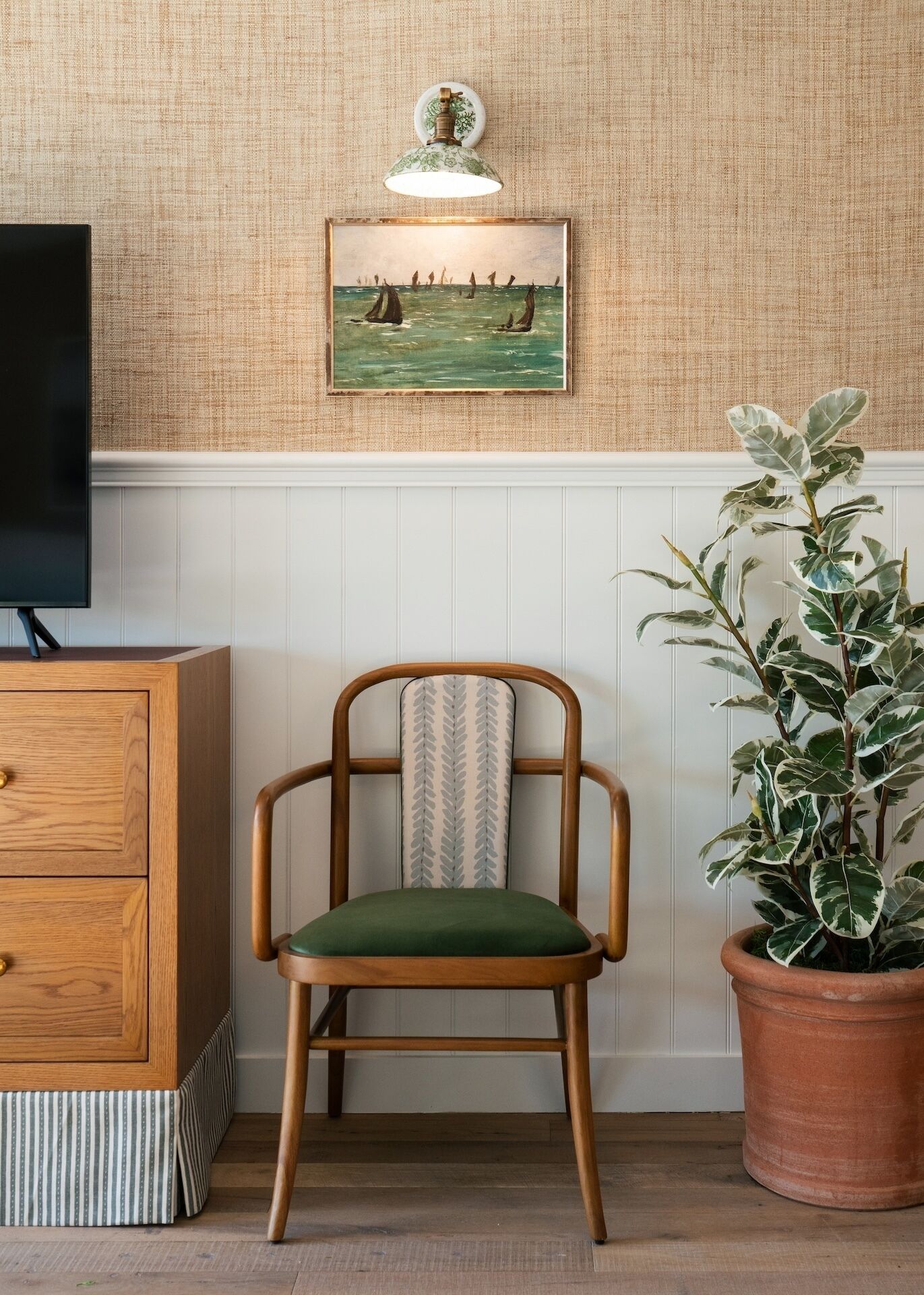 A room with a wooden chair with a patterned cushion, a potted plant, a wall-mounted light fixture, and a framed painting above a white paneled wall.